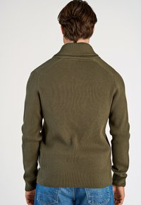 Olijfgroen geribbelde breisweater met een hoge kraag, lange mouwen en zijsplitten, gecombineerd met blauwe denim jeans.