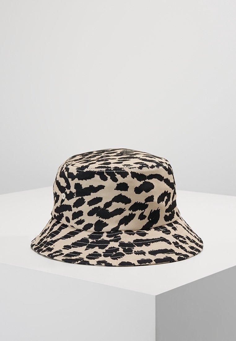 Chapeau bob beige avec motif léopard noir posé sur une surface blanche sur fond gris clair uni.