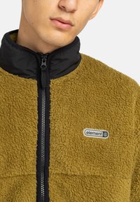 Fleecejack in mosterdgeel met een zwarte opstaande kraag. Voorzien van een rits en een logo-opdruk aan de linkerkant. Zachte textuur.
