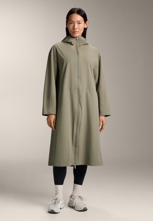 Manteau long vert olive avec fermeture éclair frontale, capuche et manches longues, porté sur des leggings noirs et des sneakers gris. Texture de tissu lisse.