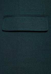 Ciemnozielona tkanina dopasowana o subtelnej fakturze, z prostokątną kieszenią u góry, podkreślająca czyste linie i zorganizowany design.