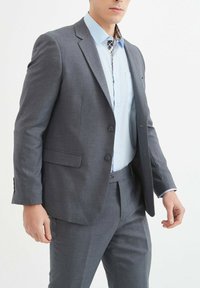 Veste de costume grise ajustée à deux boutons et à revers, associée à une chemise bleu clair avec un col à carreaux et un détail de poche.