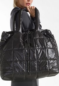 Persona che tiene una grande borsa a tracolla nera con imbottitura e manici imbottiti, indossando un indumento a maniche lunghe grigio scuro, labbra leggermente socchiuse.