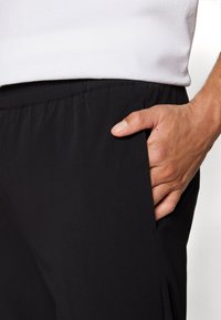 Zwarte sportieve broek met een soepele stofstructuur, een elastische tailleband en een zijzak met een hand erin gestoken.
