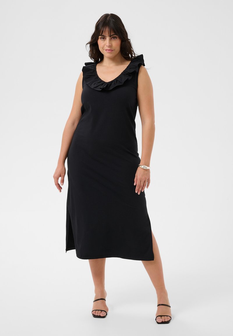 Femme debout, portant une robe midi noire sans manches avec un col en V volanté et des fentes latérales, associée à des talons ouverts noirs et un bracelet argenté.