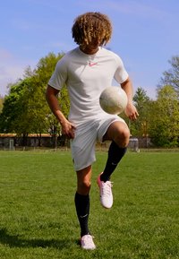 Fehér sport póló rózsaszín Nike logóval, fehér rövidnadrág, fekete zoknik és rózsaszín focicipők. A játékos egy térdén egy focilabdát egyensúlyoz.