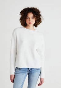 White ribbed knit sweater with a round neckline and long sleeves, paired with light blue denim jeans. Smooth texture, casual fit.