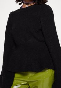 Schwarzer gestrickter Pullover mit langen Ärmeln und einem Peplum-Design, das eine weiche Textur aufweist. Getragen über glänzenden grünen Hosen.