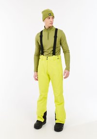Snowboardoutfit met een lichtgroen longsleeve bovenstuk en felgele broek met zwarte bretels. Zwarte boots maken de look af.