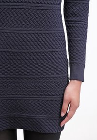 Gros plan sur une robe pull en tricot foncé avec des motifs texturés variés, portée par une personne avec une main détendue le long du corps.