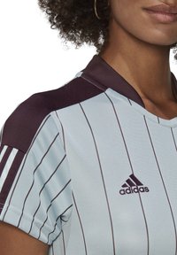 Maillot de sport bleu clair avec des rayures verticales foncées, un col sombre et le logo Adidas sur la poitrine. Le tissu est respirant et texturé.