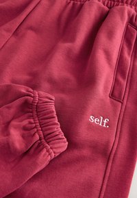 Pantaloni della tuta rosa scuro con polsini elastici e tasca laterale, con testo ricamato bianco "self." vicino alla tasca.