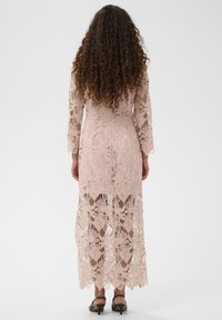 Rochie maxi din dantelă de culoare roz deschis, cu mâneci lungi, modele florale și un tiv ondulat. Țesătura texturată este semi-transparentă.