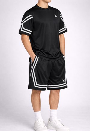 Uomo che indossa una maglietta sportiva nera e pantaloncini con strisce geometriche bianche, calzini bianchi e scarpe da ginnastica bianche, in piedi con le mani in tasca.