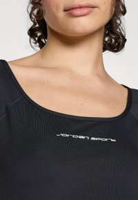 Svart sportstopp med rundad halsringning, med en mjuk textur och texten "Jordan Sport" i vitt över bröstet.