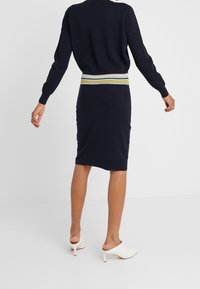 Vestito a maglia blu navy dalla forma aderente, con una fascia elastica a righe gialla, bianca e blu in vita. Maniche lunghe e lunghezza fino a metà polpaccio.