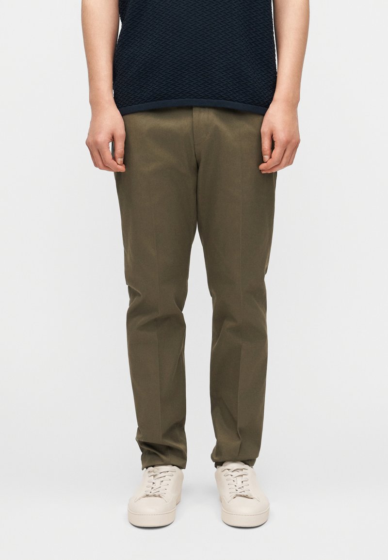 Άτομο που φοράει παντελόνι σε ευθε ία γραμμή, ελιάς πράσινο, off-white αθλητικά παπούτσια και υφασμάτινο κοντομάνικο τοπ σε σκούρο ναυτικό χρώμα, στέκεται σε ανοιχτό φόντο.