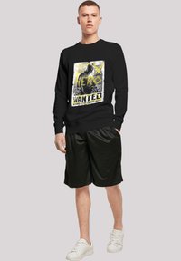 Sudadera negra con estampado gráfico que presenta texto en amarillo y blanco, combinada con pantalones cortos negros holgados y zapatillas blancas. Textura suave, ajuste casual.