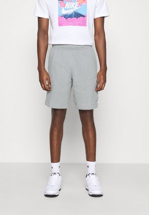 Mężczyzna ubrany w biały t-shirt z grafiką Nike, szare spodenki z elastycznym paskiem i boczną kieszenią, białe skarpetki Nike i białe sneakersy, stojący przy białej ścianie.