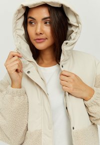 Beige padded jacket with a fur-like texture on sleeves. Features a hood and button closure, offering a warm and casual design.