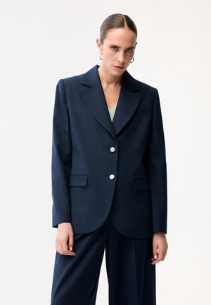 Kvinde med tilbageglattet hår iført en marineblå dragt med sølvknapper og øreringe i hulringe, stående foran en ensfarvet hvid baggrund.