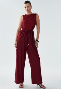 Rdečkast ogrinjalo z naborkastim zgornjim delom, širokimi hlačnicami in pasom z zavezovanjem. Ima teksturirano tkanino in brezrokaven dizajn. Črne sandale so obute.