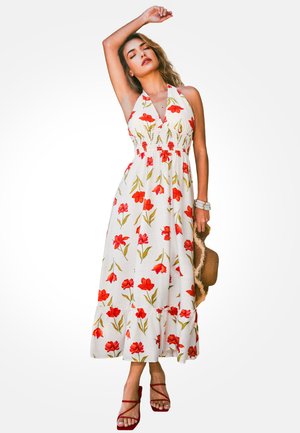 Femme portant une robe blanche à col halter avec imprimé floral rouge, talons rouges à brides, tenant un chapeau à large bord marron, posant avec un bras levé.