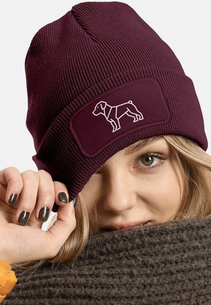 Maroon knit beanie with ribbed texture and a rectangular patch featuring a white outline of a dog. Hand touching the brim.
