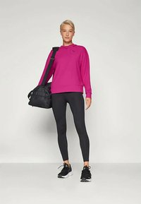 Ung kvinne i magenta sweatshirt, svarte leggings og joggesko som holder en svart treningsbag over skulderen, stående mot en ensfarget bakgrunn.