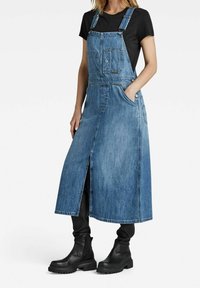 Džínová šatová šaty, světle modré, s nastavitelnými ramínky, přední kapsou, bočními kapsami a předním rozparkem. Na sobě má černý top a kalhoty.