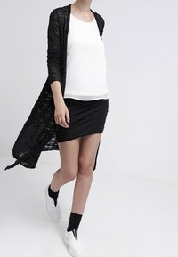 Žena jde oblečená v bílém topu bez rukávů, černé mini sukni, dlouhém černém kardiganu, černých ponožkách a bílých slip-on botách.