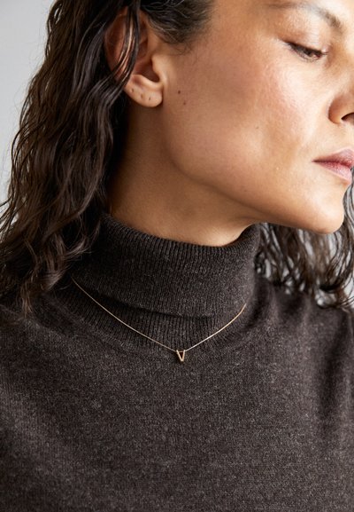 Gold pendant necklace with a 'V' shape, worn over a dark brown, textured turtleneck sweater. Hair is wavy and shoulder-length.