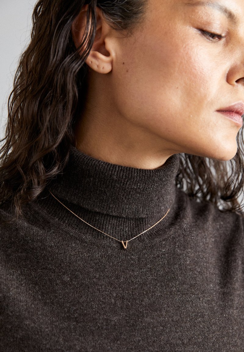 Kuldpendantkaelake, millel on 'V' kuju, kantud tumepruunist tekstuuriga kõrge kaelusega kampsunist. Juuksed on lainelised ja õlgadesse ulatuvad.