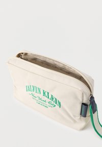 Μπεζ θήκη Calvin Klein με πράσινο κεντημένο λογότυπο, φερμουάρ στην κορυφή και υφασμάτινο λουράκι καρπού σε πράσινο και μπεζ χρώμα.