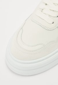 Baskets blanches présentant un mélange de cuir lisse et de daim. Design arrondi avec des détails de couture et une semelle en caoutchouc texturé.