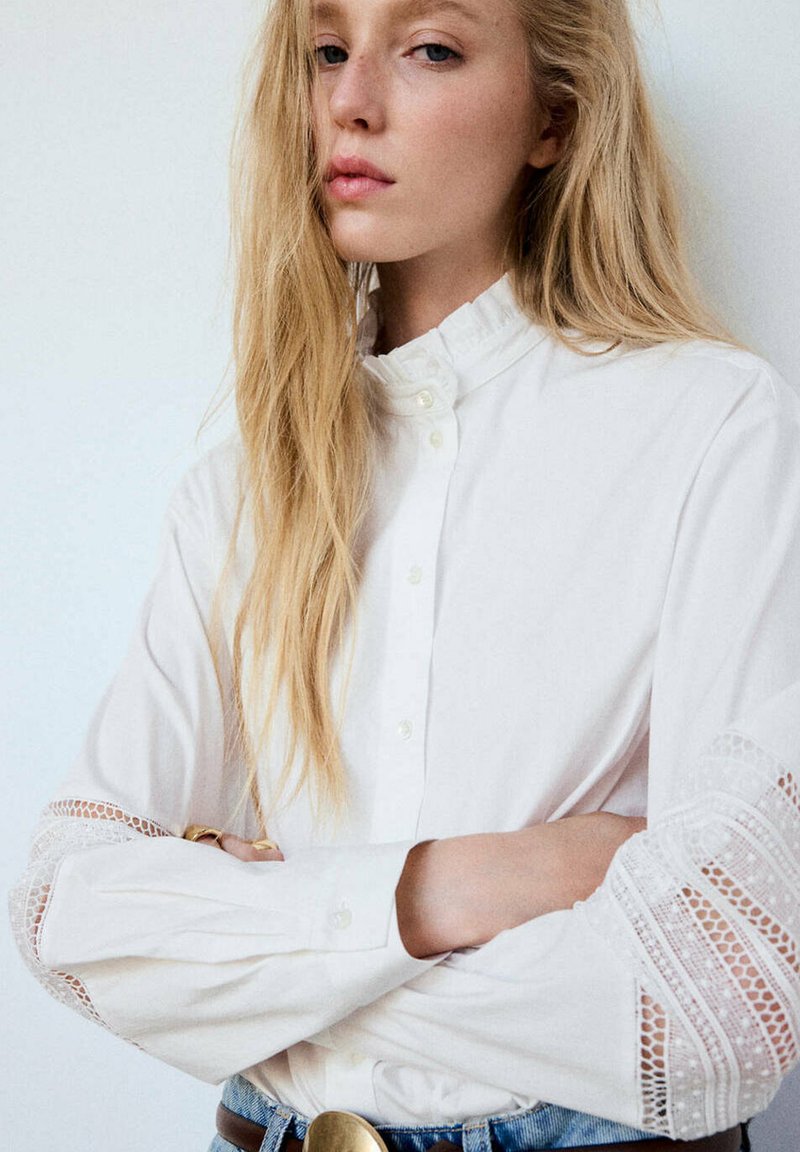Femme blonde aux longs cheveux portant un chemisier blanc avec des détails en dentelle sur les manches, les bras croisés, sur un fond clair uni.