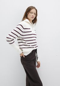 Weißes geripptes Sweatshirt mit kastanienbraunen Streifen und einem halben Reißverschluss-Kragen. In Kombination mit dunkelbraunen Hosen steht das Modell seitlich.