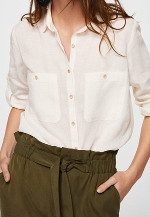 Kvinde iført en løs hvid skjorte med knapper, to brystlommer og opkørte ærmer, kombineret med højtaljede grønne bukser med bælte bundet i taljen.