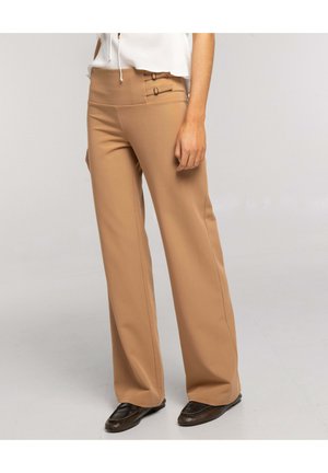 Mujer con pantalones beige de talle alto y pierna ancha, con detalles de doble hebilla, combinados con una blusa blanca y mocasines marrón oscuro.
