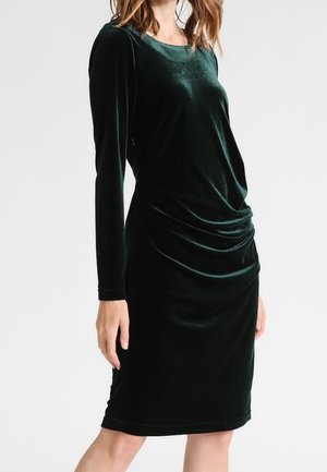 Femme portant une robe en velours vert foncé, cintrée, à manches longues, longueur genou, avec un léger froncé à la taille.
