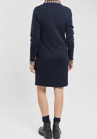 Femme debout, de dos, portant une robe bleu marine à manches longues par-dessus une chemise à carreaux, des chaussettes bleu marine et des bottines noires sur un fond uni.