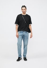 Zwart T-shirt met korte mouwen en witte bies, lichtblauwe spijkerbroek en zwarte sneakers met witte accenten. Model houdt een zwarte crossbodytas vast.