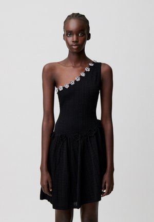 Donna che indossa un vestito nero monospalla con dettagli floreali bianchi lungo lo scollo e dettagli floreali neri in vita.