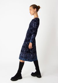 Robe navy à manches longues avec un motif floral en bleu clair. Elle présente une jupe plis et des accents côtelés. Associée à des bottes noires hauteur genou.