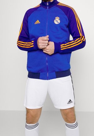 Homme portant une veste de survêtement Adidas bleue et violette avec des rayures orange et le logo du Real Madrid, un short blanc Adidas et des chaussettes blanches rayées.