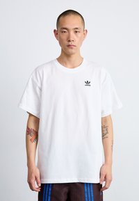 Tricou alb din bumbac, cu guler rotund, mâneci scurte și un mic logo negru adidas pe pieptul stâng. Purtați-l cu pantaloni scurți cu motive.