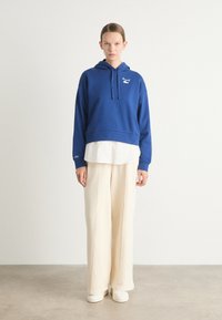 Felpa cropped blu con cappuccio regolabile, logo bianco, indossata sopra una maglietta bianca, abbinata a pantaloni larghi crema e calzature bianche.
