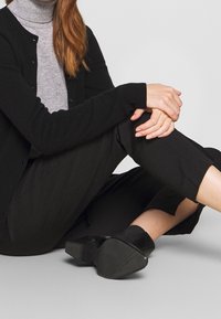 Person som sitter i skräddarställning, iklädd svarta byxor, svarta stövletter med klack, svart cardigan och grå polo-tröja.