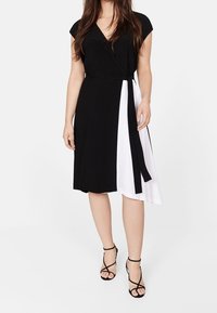 Schwarz-weißes Wickelkleid mit kurzen Ärmeln, das eine gebundene Taille, einen knielangen Saum und eine glatte Stofftextur aufweist. Schwarze Riemchenheels.