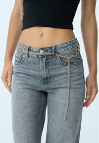 Goldene Kettengürtel mit runden Gliedern und einem hängenden Kettendetail, getragen über hochtaillierten grauen Denim-Jeans. Glatte Textur und metallischer Glanz.